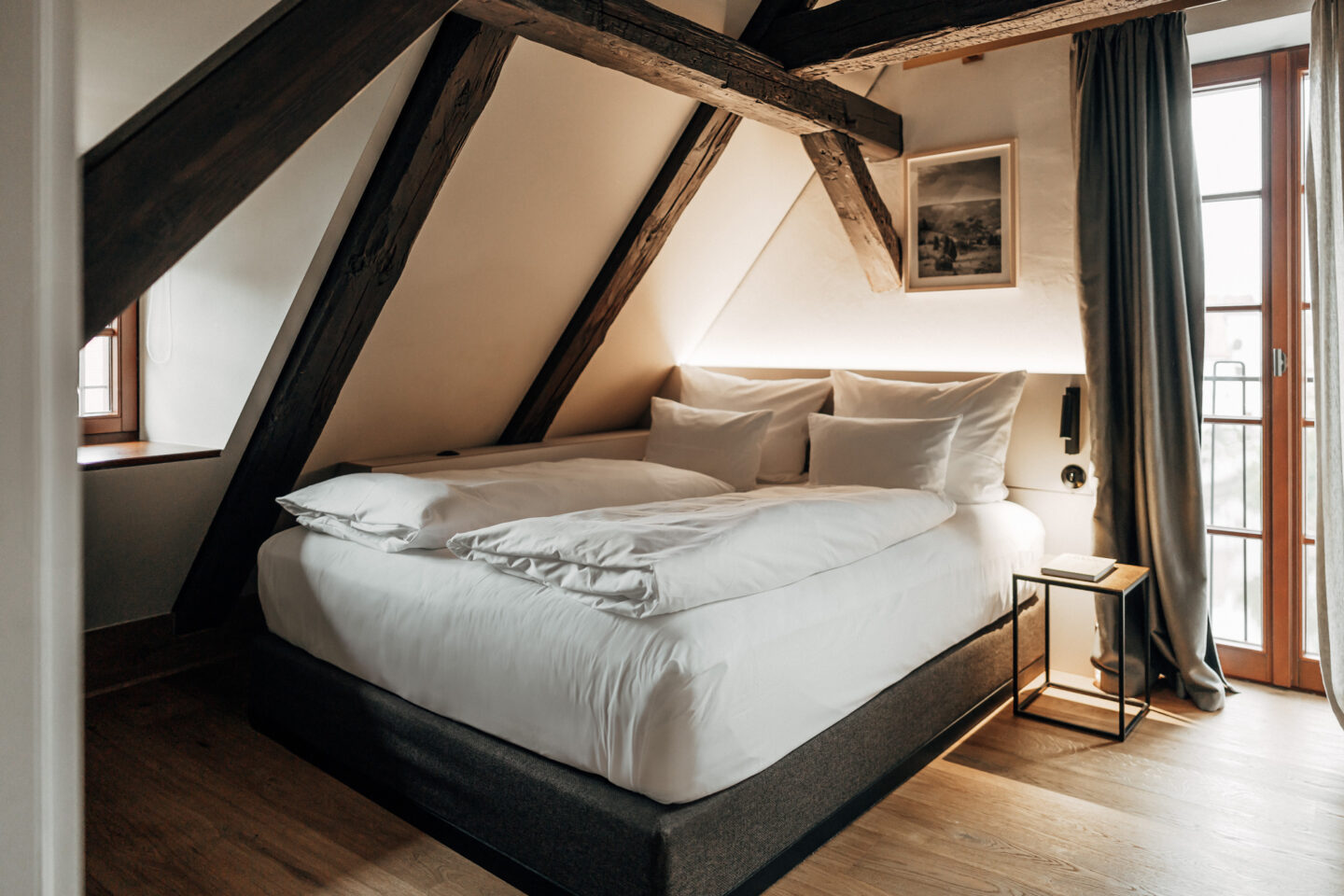 Cozy attic bedroom with a large bed, wooden beams, and a small bedside table, featuring soft lighting and curtains.