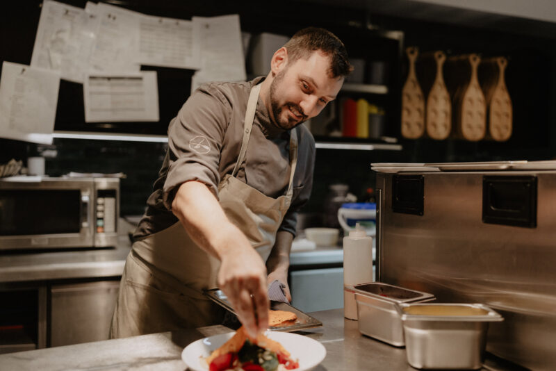 Chef plating a colorful dish with fruits in a modern kitchen, smiling while working at a counter.