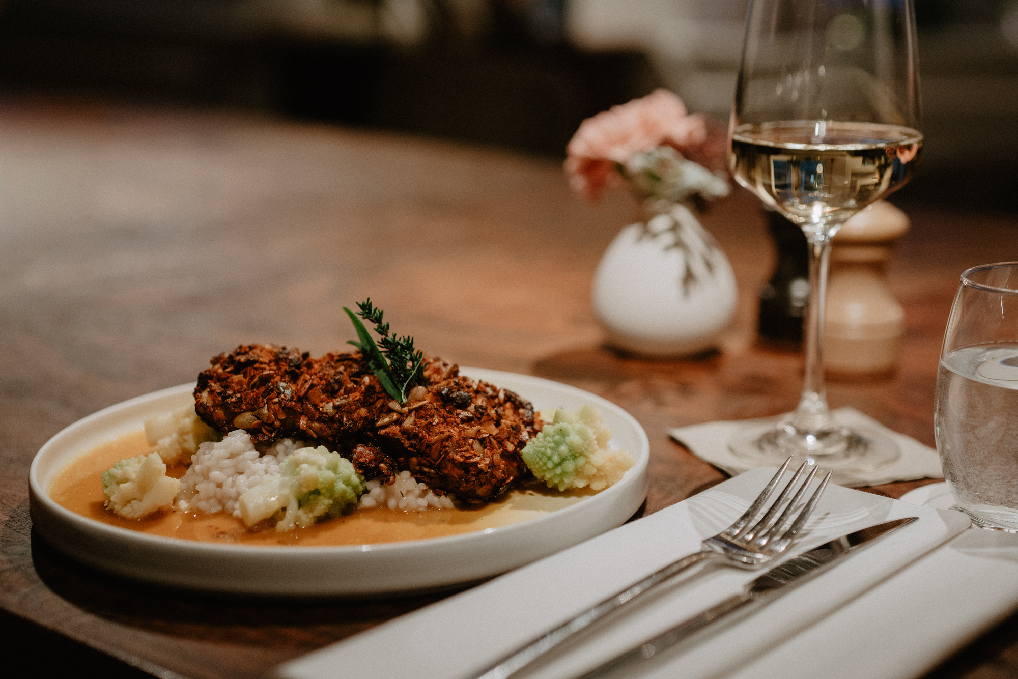 Crispy vegetable patties on a bed of rice, garnished with herbs, accompanied by a glass of white wine and a flower vase.