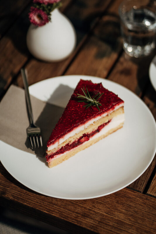 Slice of layered cake with raspberry topping on a white plate, fork beside it, set on a wooden table.