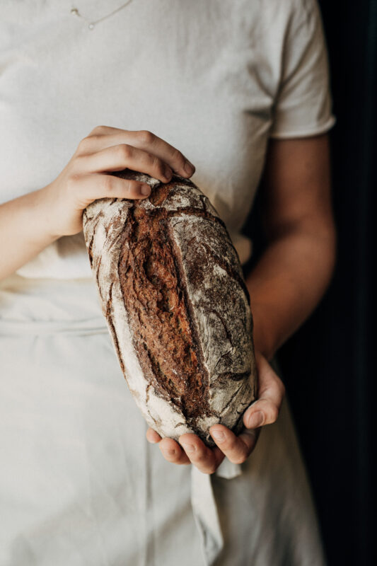 A person holding a rustic loaf of bread, wearing a light-colored shirt and apron, against a dark background.