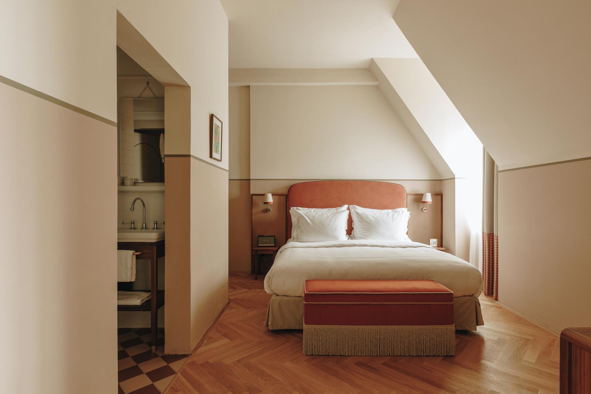 Modern hotel room with a sloped ceiling, featuring a double bed, orange accents, and a cozy seating area.