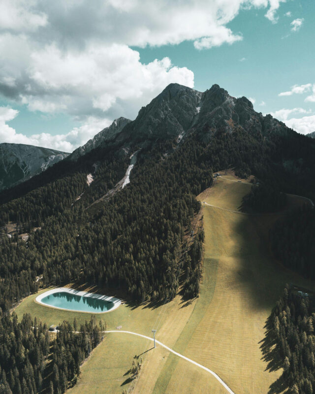 Aerial view of Castel Badia, featuring a mountain backdrop, lush green fields, and a small pond surrounded by trees.