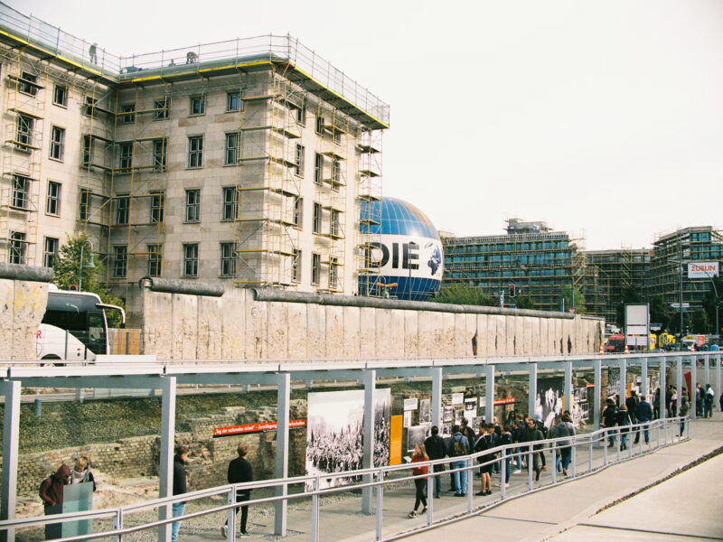 Visitors line up at the Berlin Wall memorial, with construction scaffolding and a large globe in the background.