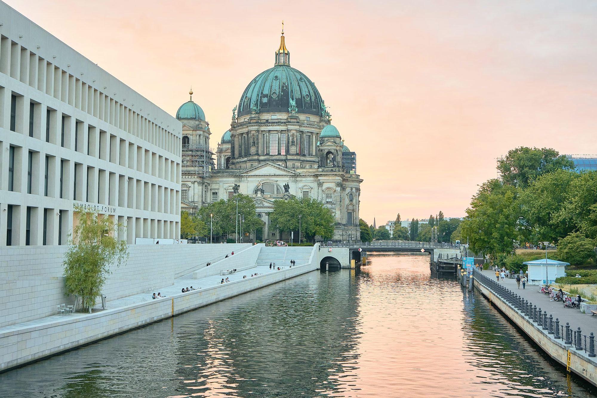 Berlin Cathedral viewed from a riverbank at sunset, with modern architecture on the left and lush greenery along the water.