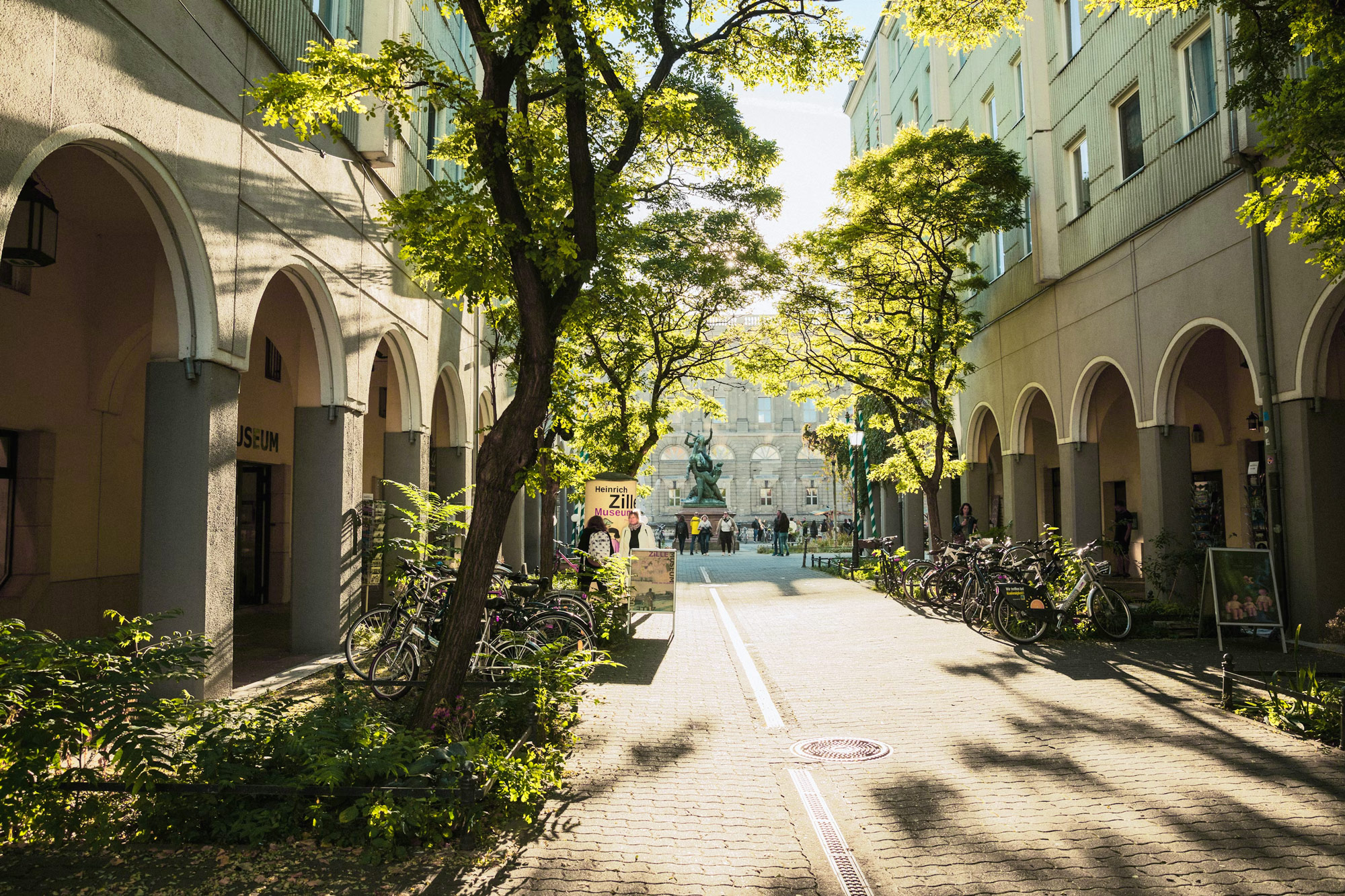 Sunlit alleyway lined with trees and bicycles, leading to a square with a fountain and people in the background.