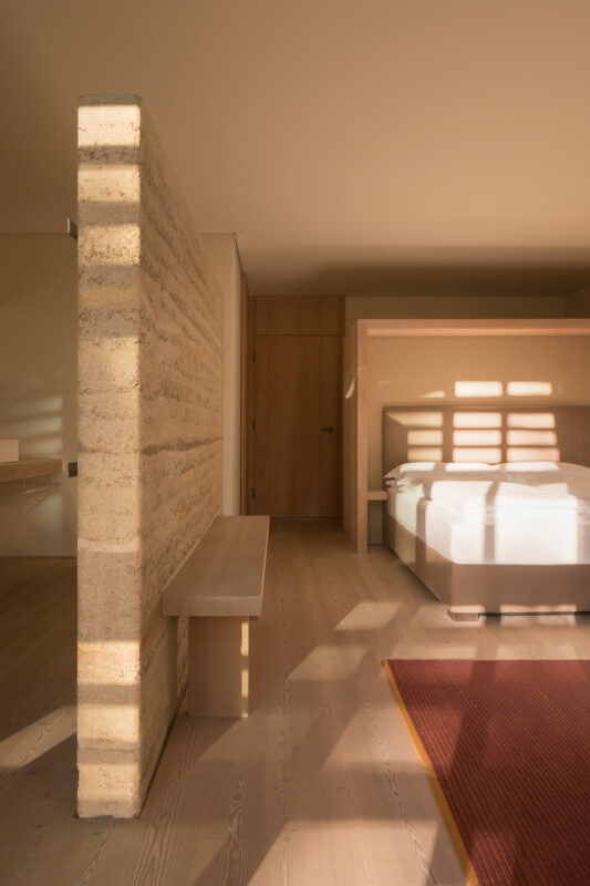 Modern bedroom with a textured wall, wooden bench, and soft lighting casting shadows on the floor.