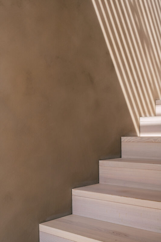 Light wooden stairs ascend against a textured brown wall, with shadows creating a striped pattern on the surface.