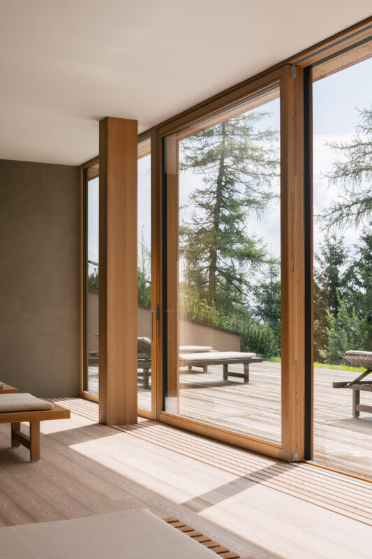 Bright, modern relaxation room with large glass windows, wooden accents, and a view of outdoor seating and trees.