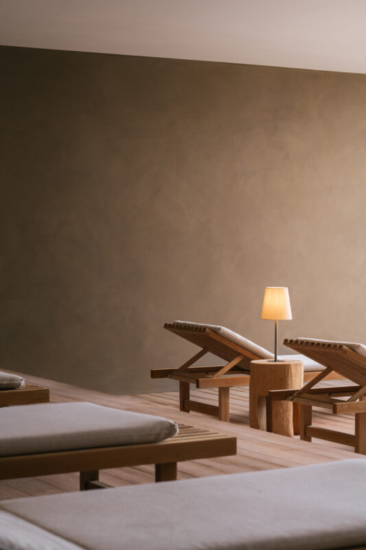 Relaxation room featuring wooden lounge chairs, a small round table, and a lamp against a textured brown wall.