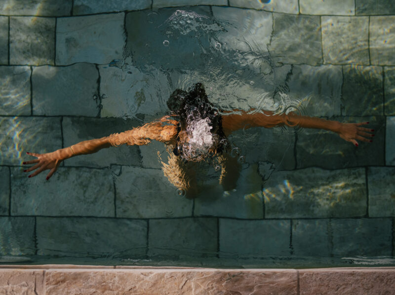 Aerial view of a person with arms outstretched in a tranquil spa pool, surrounded by stone tiles.