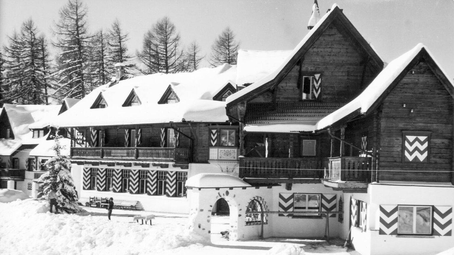 Historic wooden chalet with snow-covered roof and zigzag patterns, surrounded by snow and pine trees.