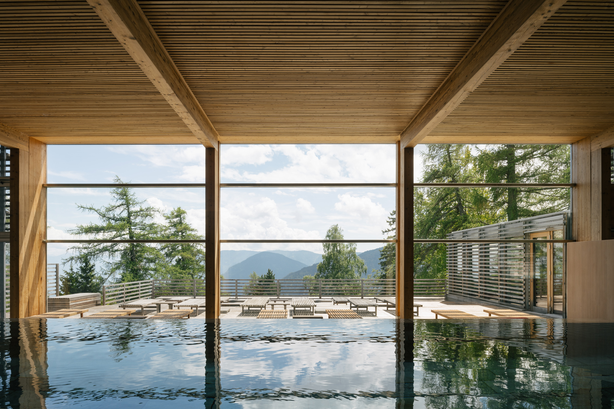 Indoor swimming pool with wooden beams, overlooking a scenic view of trees and mountains through large glass windows.