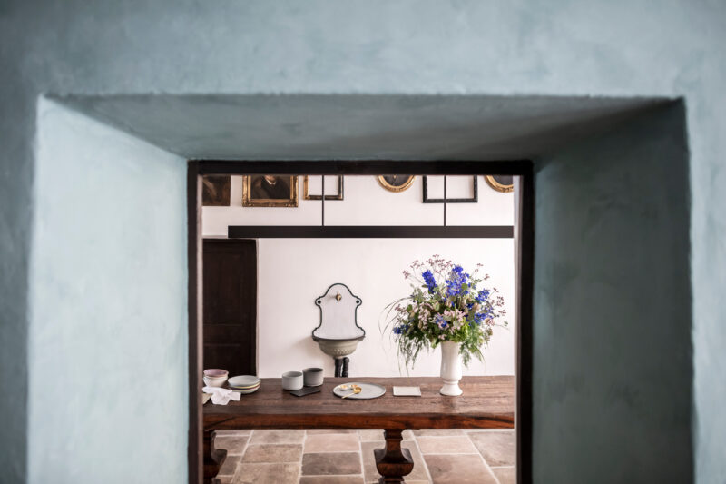 View through a stone archway into a room with a wooden table set with dishes and a vase of blue flowers.