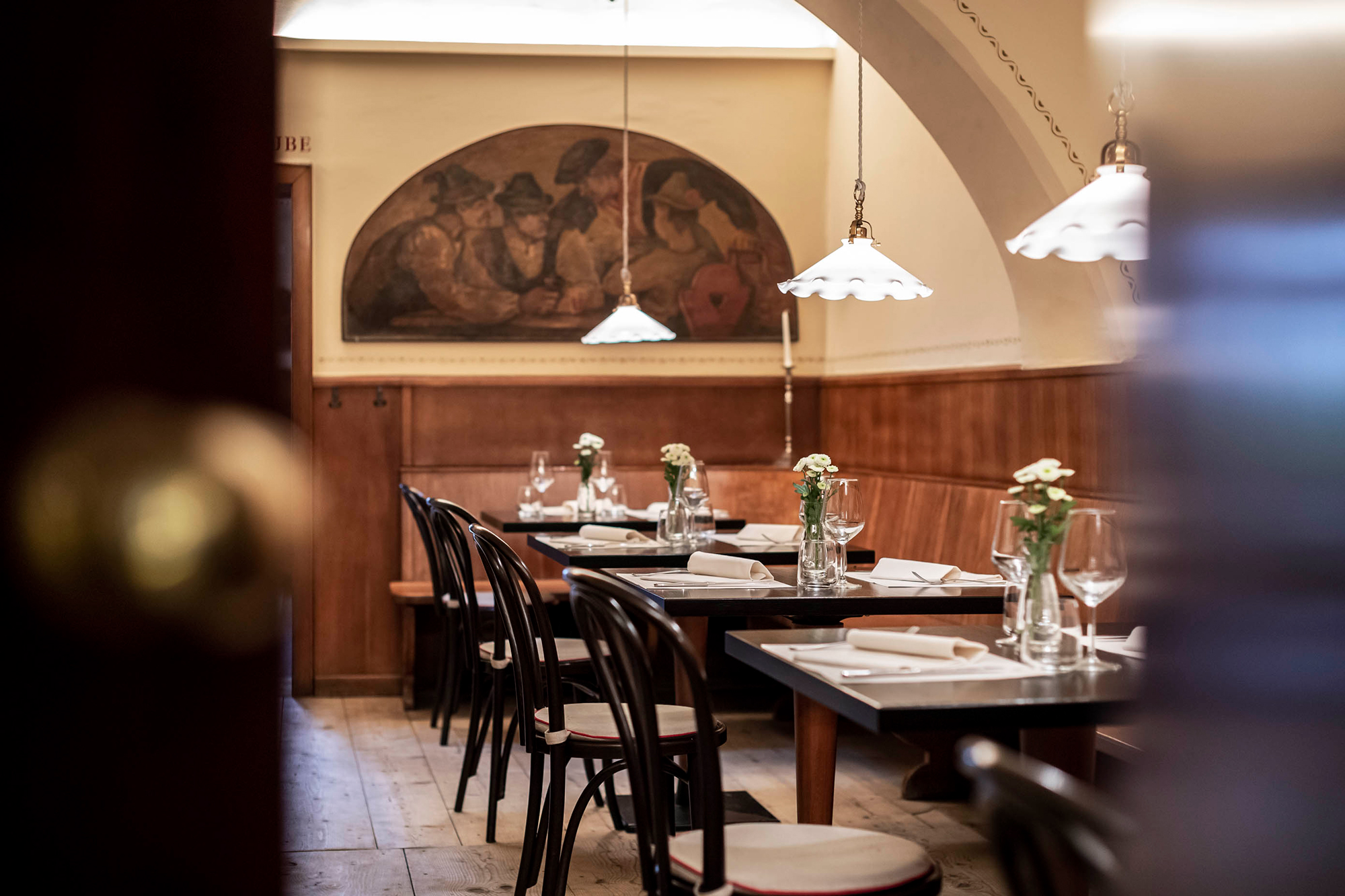 Cozy restaurant interior with wooden tables, chairs, and floral centerpieces, featuring a mural on the wall.