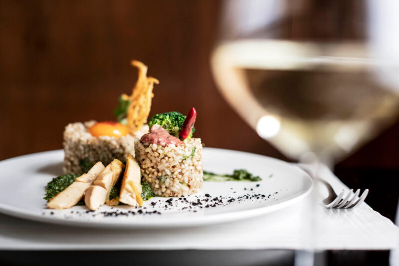 Gourmet dish featuring bulgur, chicken, and garnishes, with a glass of white wine in the foreground.