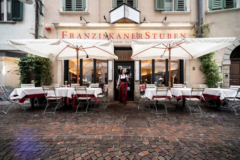 Outdoor dining area of Franziskaner Stuben with tables under umbrellas and a woman in traditional attire.