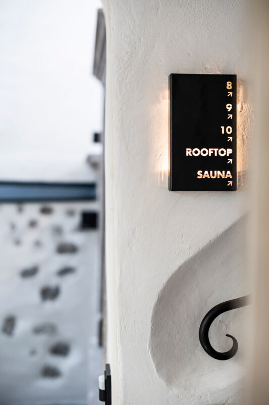 Sign indicating "Rooftop Sauna" mounted on a wall, with a blurred stone wall in the background.