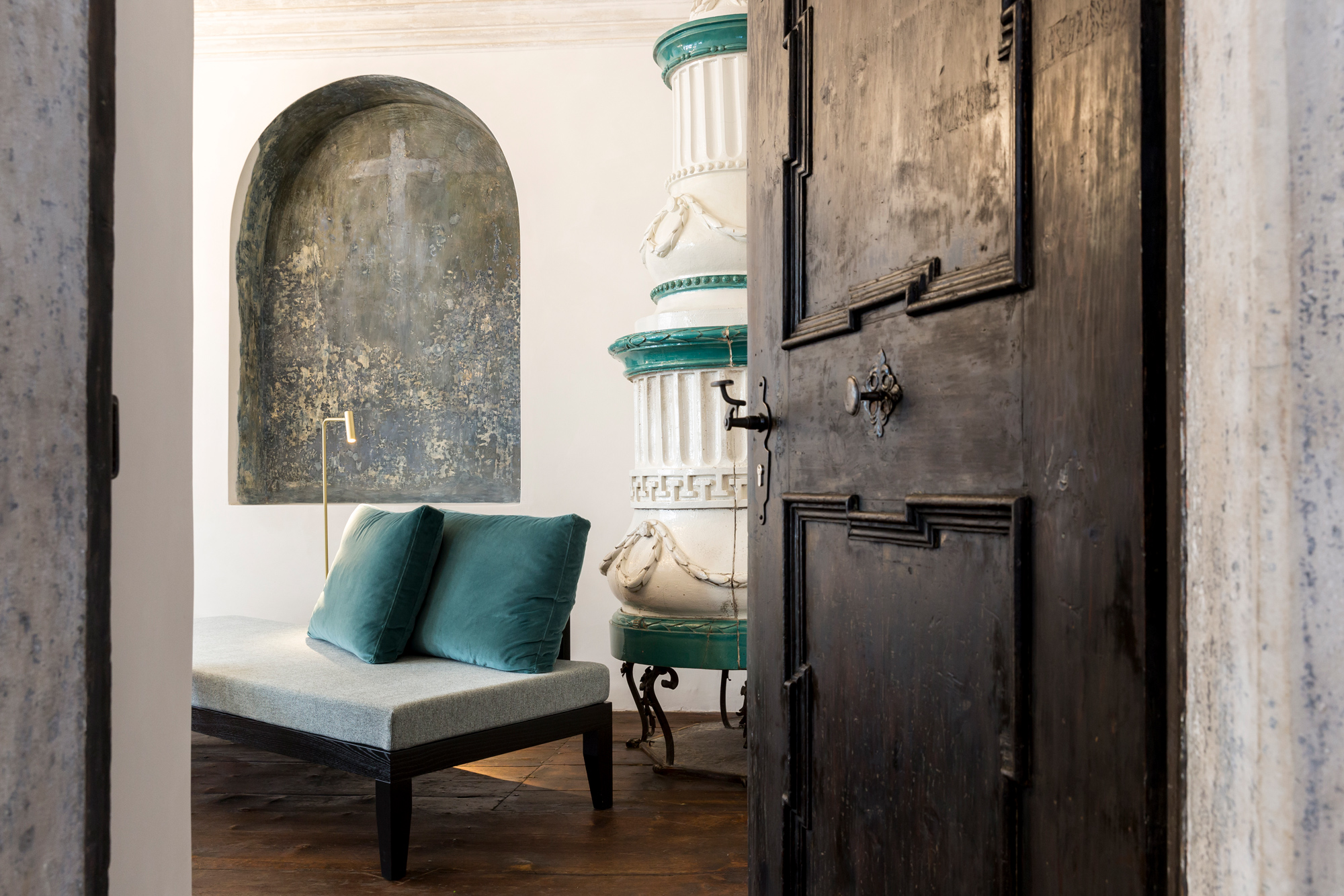 View into a stylish junior suite featuring a wooden door, a chaise lounge with teal cushions, and decorative architectural...