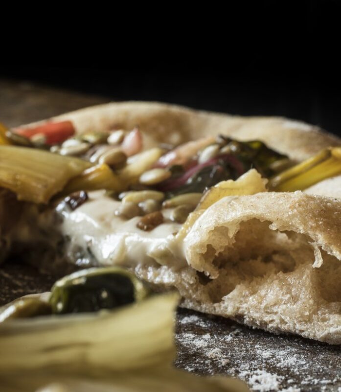 Close-up of a rustic pizza with toppings of greens, seeds, and vegetables, partially eaten, on a wooden surface.
