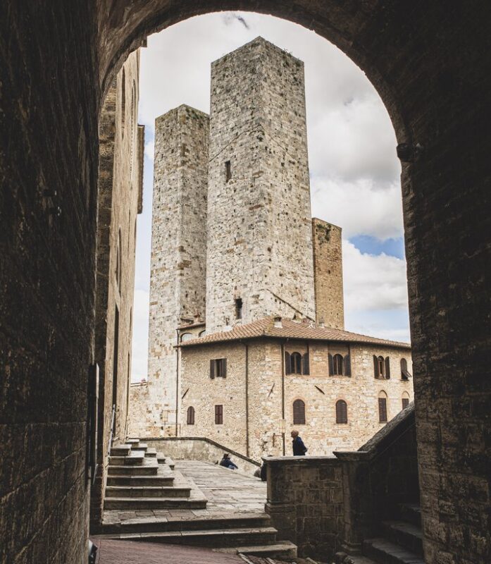 View through an archway showcasing three tall stone towers and a historic building in a medieval town.