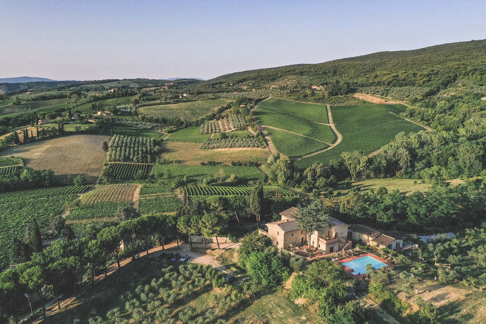 Aerial view of a Tuscan landscape featuring rolling hills, vineyards, and a rustic villa with a pool.
