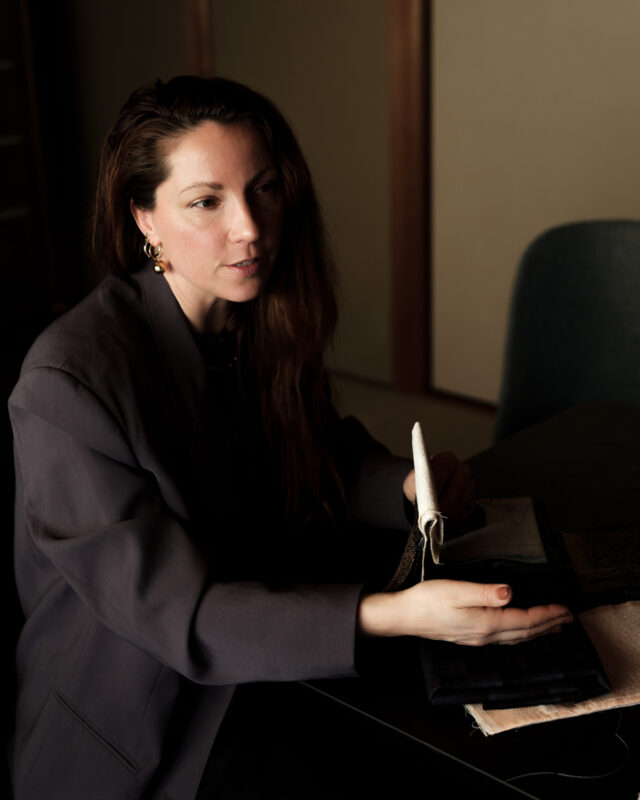 Woman with long hair in a blazer gestures while examining fabric swatches on a table.