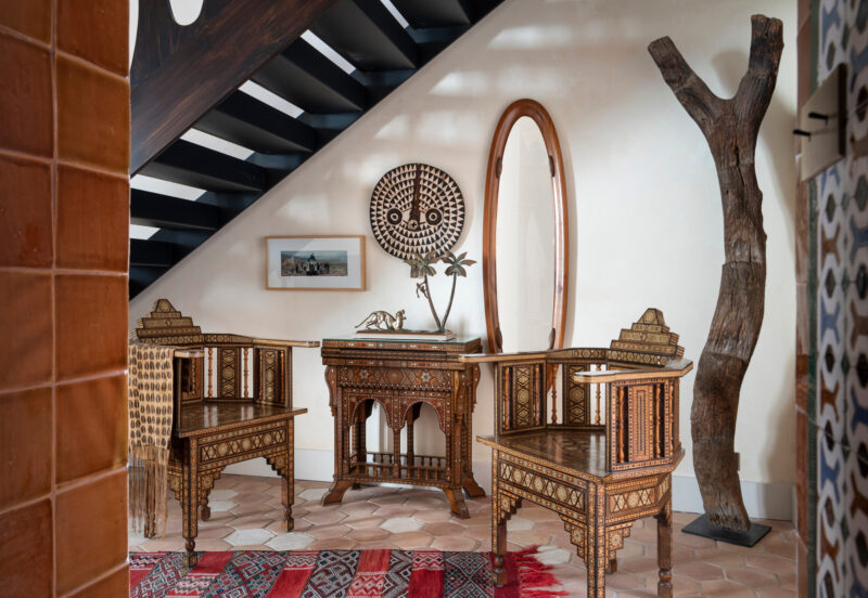 Intricately designed wooden chairs flank a decorative table under a mirror, with a staircase and a tall sculpture in the b...
