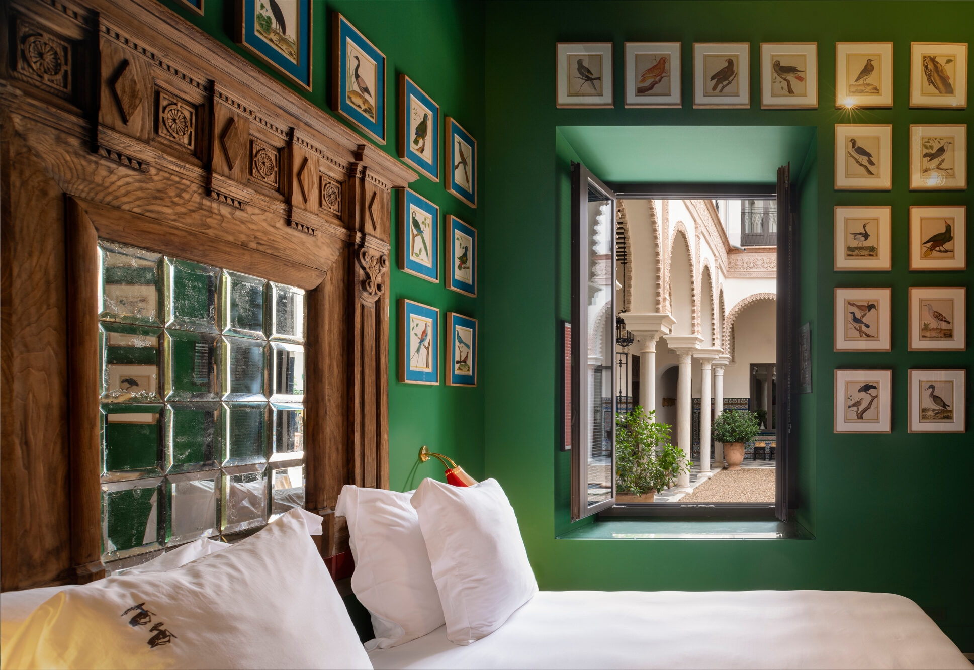 Cozy hotel room with green walls, a wooden headboard, and framed bird illustrations, overlooking a courtyard through an op...