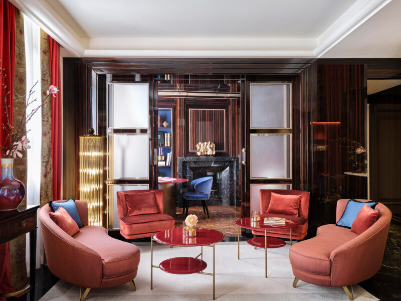 Elegant lounge area with pink velvet chairs and a round glass table, featuring a black marble fireplace and decorative acc...