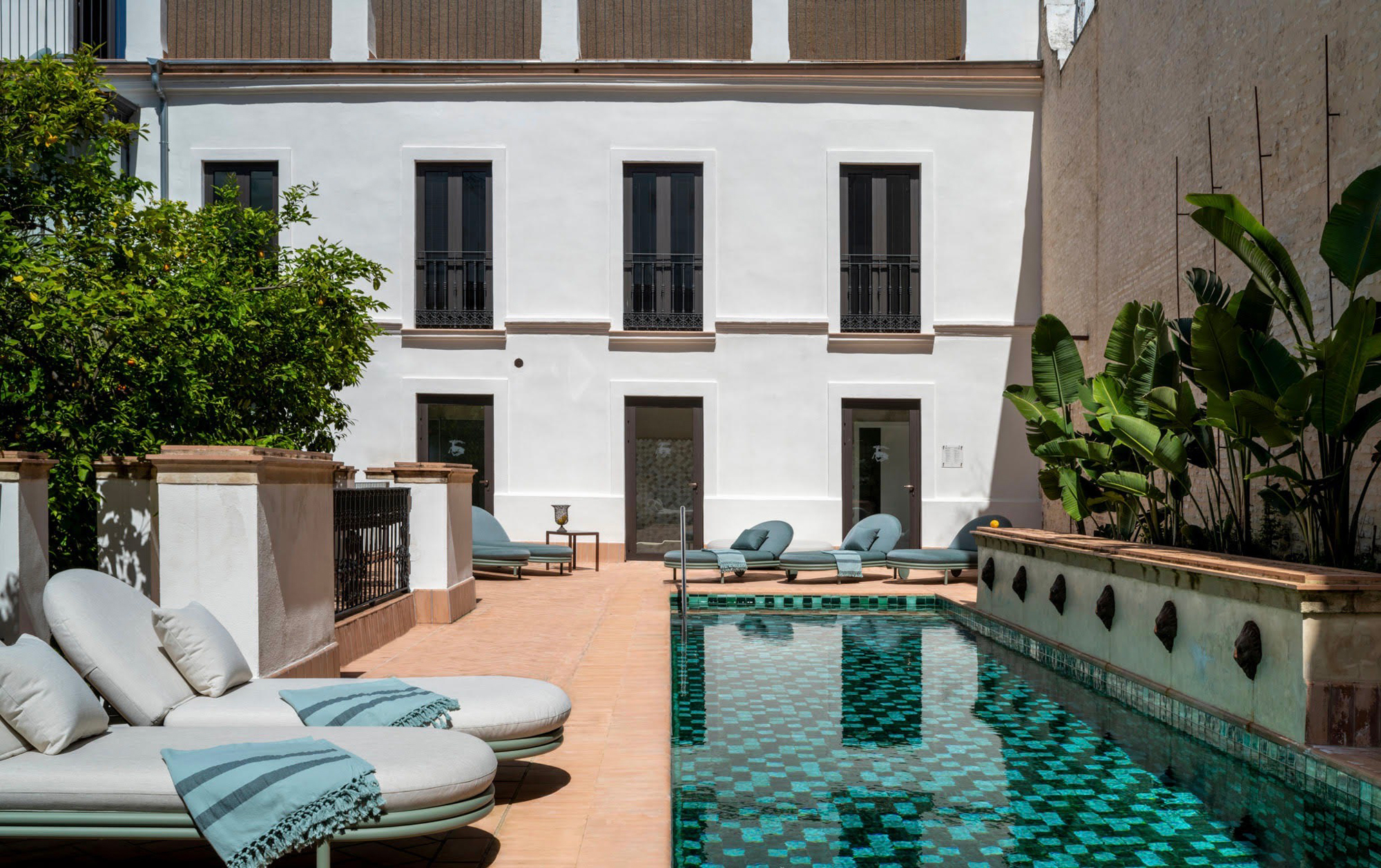 Modern hotel courtyard featuring a turquoise-tiled pool, lounge chairs, and greenery against a white building backdrop.