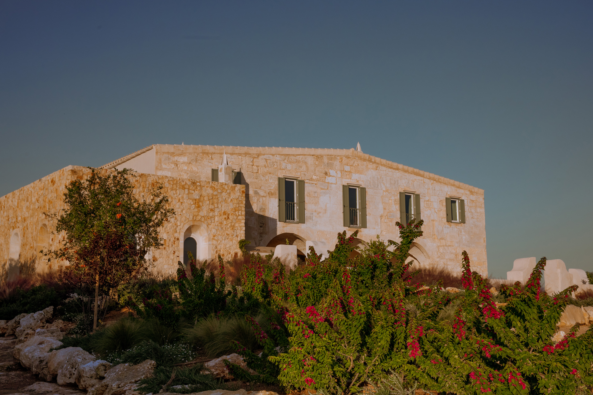 Stone building with green shutters surrounded by lush plants and flowers, set against a clear sky at Son Blanc, Menorca.