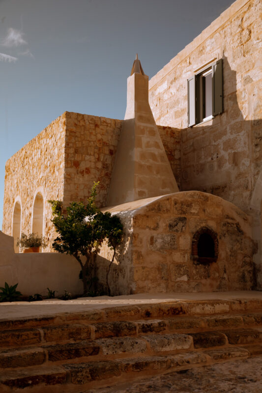 Stone building with a conical roof and arched windows, surrounded by greenery, under a clear blue sky.