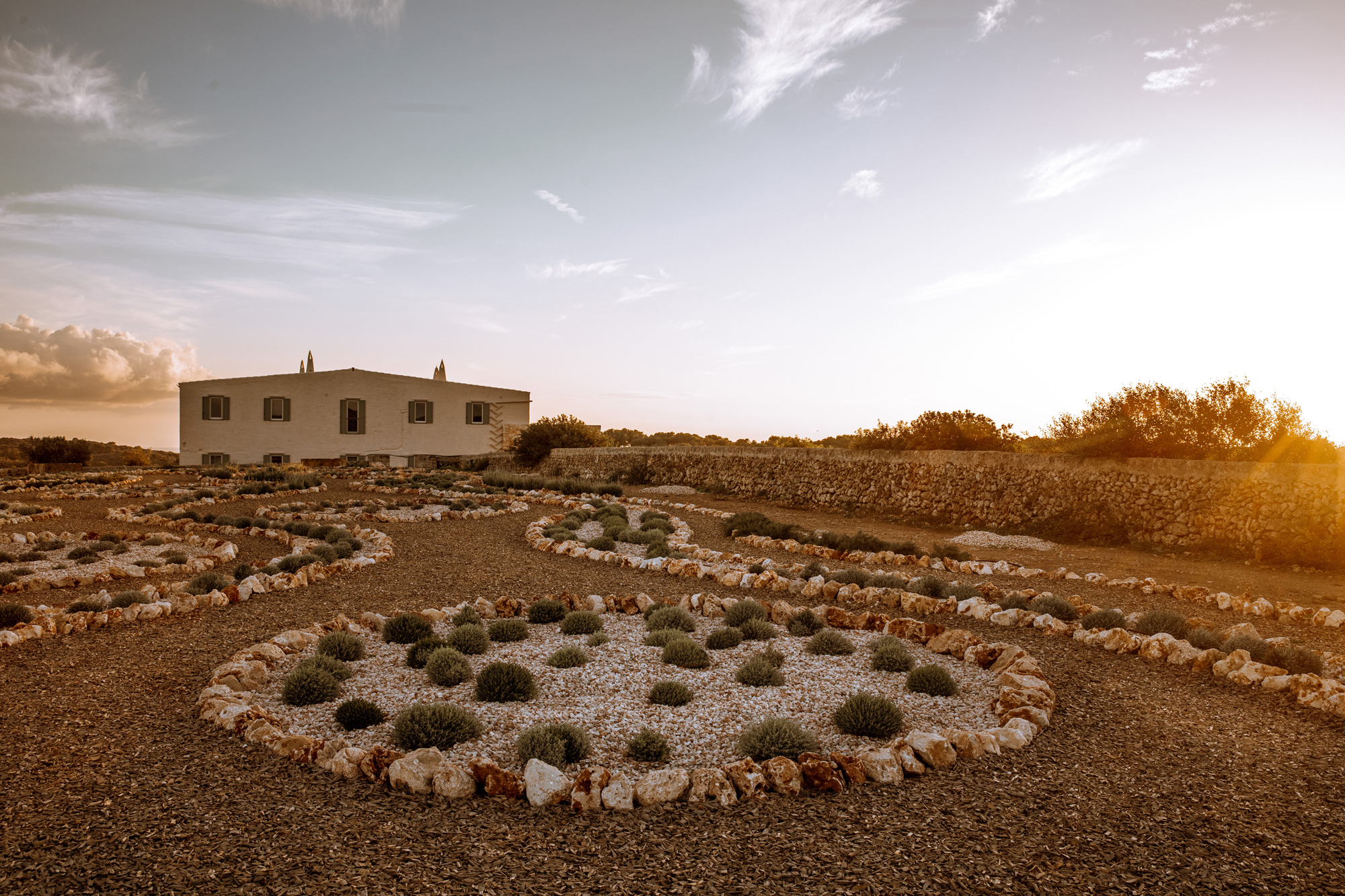 Landscape featuring a stone house surrounded by circular garden beds at sunset, with a clear sky and distant trees.