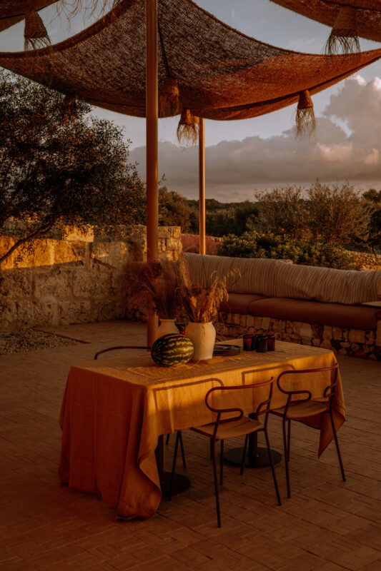 Warmly lit outdoor dining area with a yellow tablecloth, three black chairs, and decorative plants, surrounded by nature.