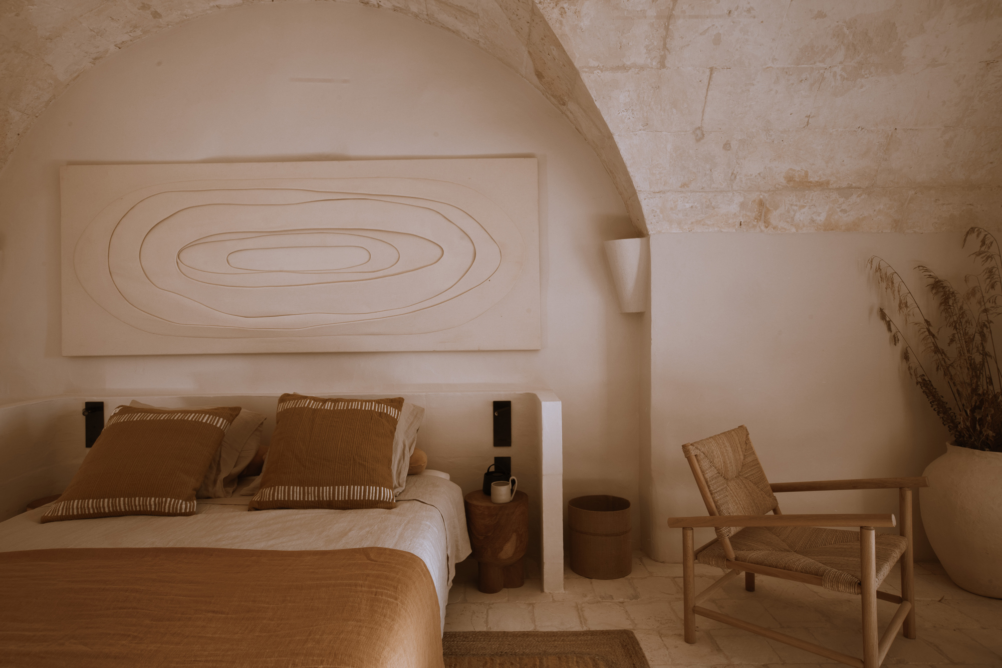 Minimalist bedroom with a curved art piece above a bed, featuring earthy tones, a wooden chair, and decorative plants.