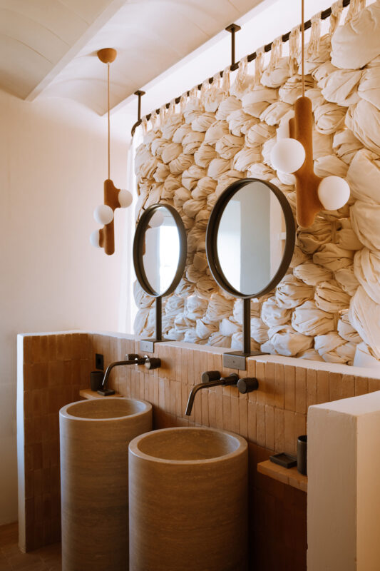 Modern bathroom featuring two round stone sinks, circular mirrors, and a textured wall with soft fabric accents.