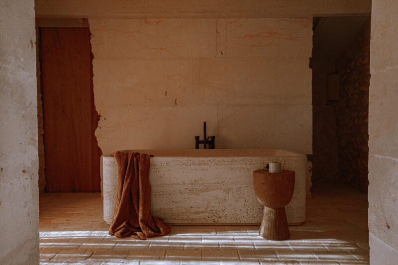 Minimalist bathroom featuring a stone bathtub, wooden stool, and a cozy blanket draped over the tub, with warm lighting.