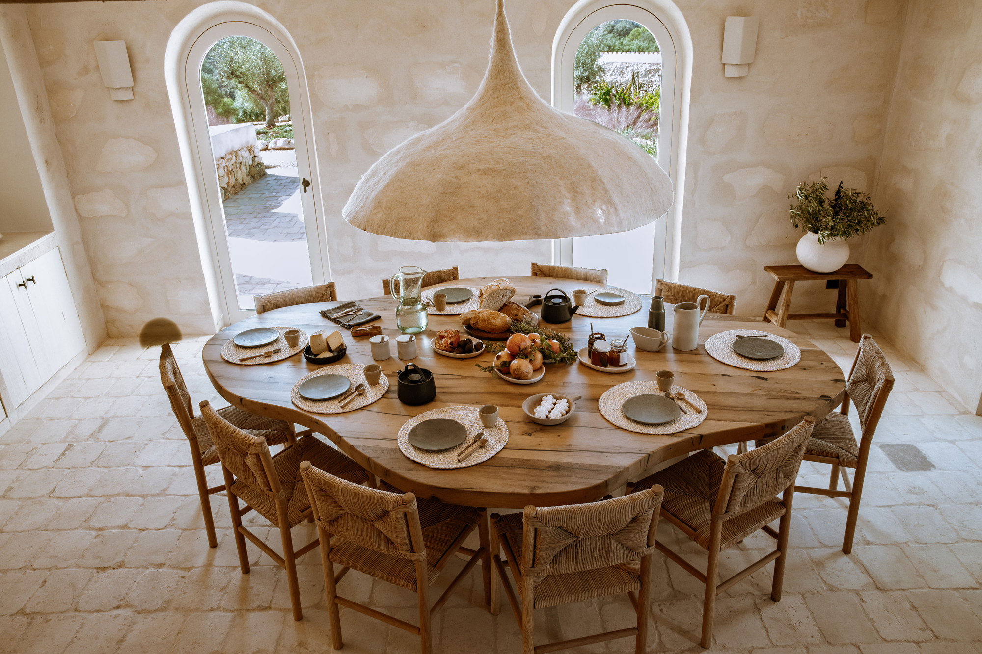 A rustic dining table set for a meal, surrounded by chairs, with a large pendant light overhead and views of a garden thro...