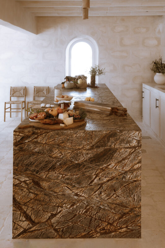 Modern kitchen featuring a textured stone island with a variety of fruits, cheeses, and a window in the background.
