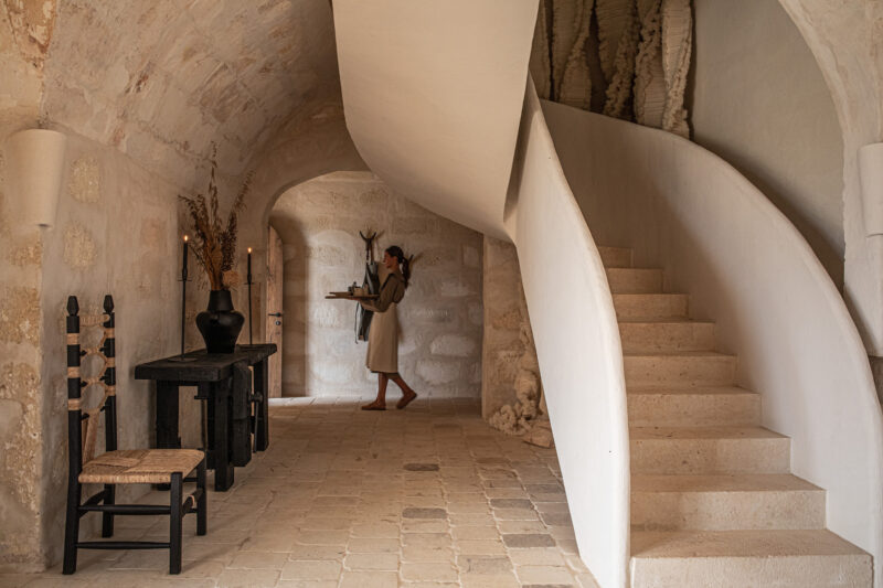 Woman carrying a tray in a minimalist interior with a curved staircase and rustic decor in Son Blanc, Menorca.
