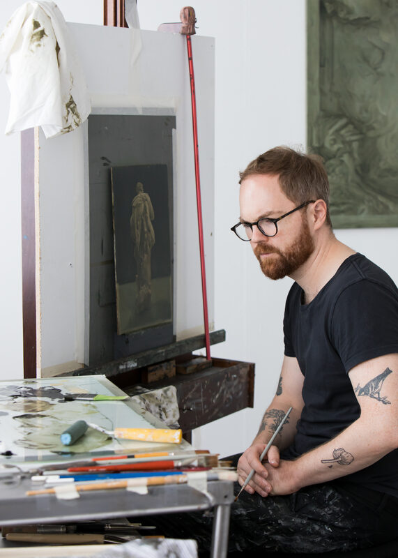 Artist Niklas Asker sits at his easel, surrounded by paints and brushes, focused on a sculpture sketch.