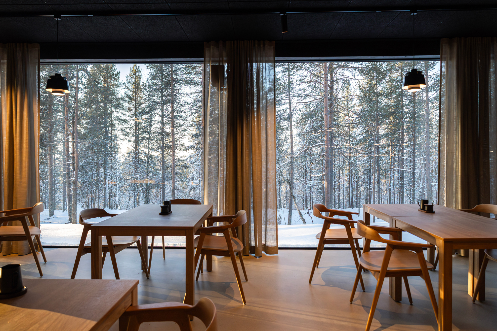 Modern dining area with wooden tables and chairs, large windows showcasing a snowy forest landscape outside.