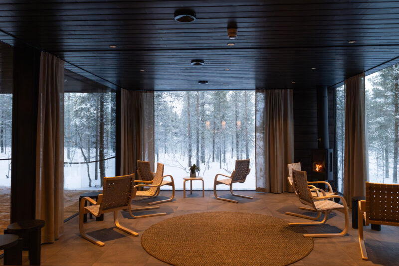 Cozy lounge area with modern chairs and a round rug, surrounded by large windows showing a snowy forest.