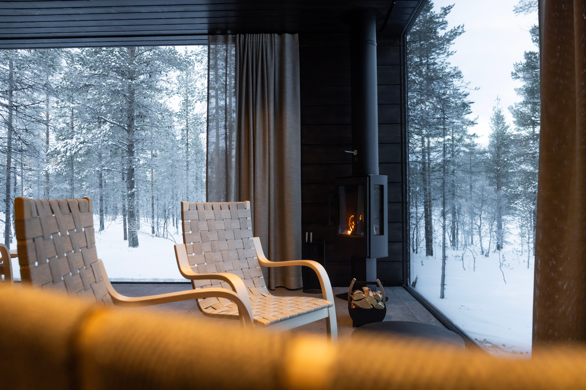 Cozy interior of Gáldu Hotel & Spa with two chairs, a fireplace, and a snowy forest view through large windows.