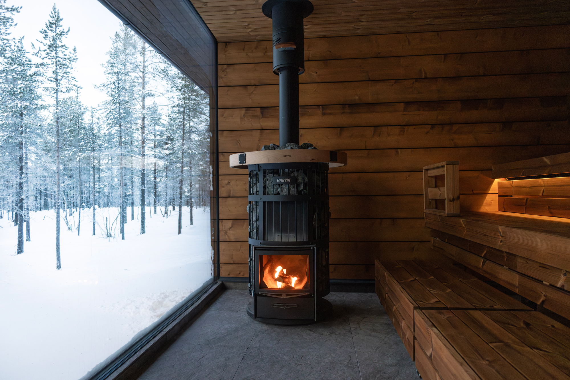 Cozy wood-burning stove in a wooden sauna, with a snowy forest visible through large glass windows.