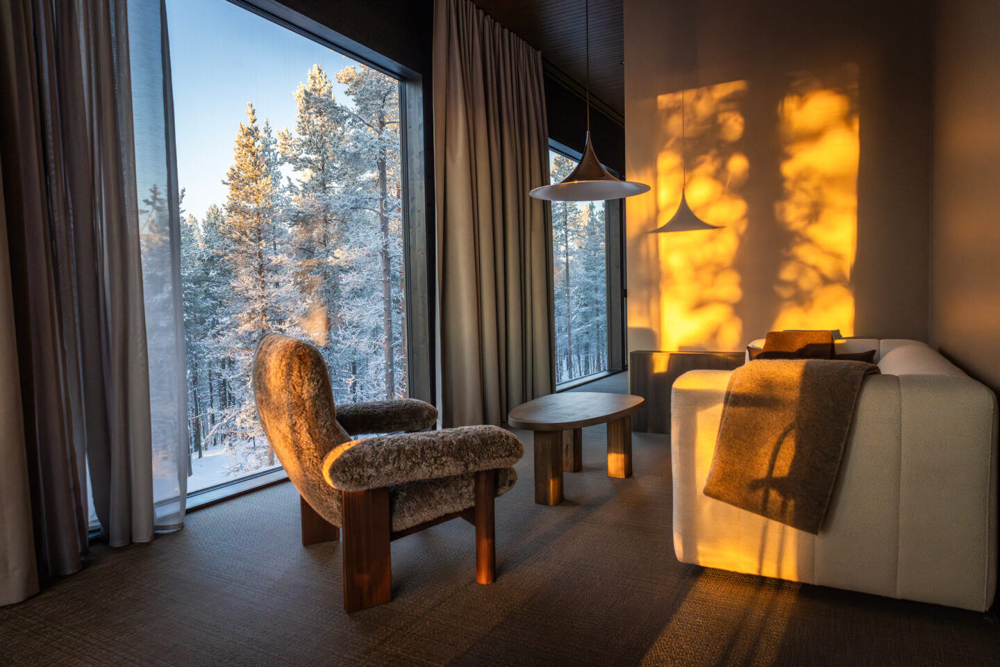 Cozy hotel room with a plush chair and a sofa, sunlight casting warm shadows on the walls, and snowy trees visible outside.