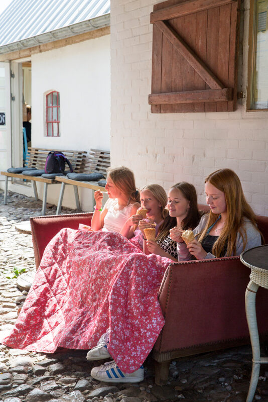Four girls sitting on a couch outdoors, wrapped in blankets, enjoying ice cream on a sunny day.