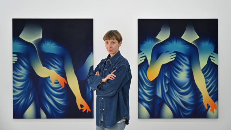 Aistė Stancikaitė stands between two colorful artworks depicting stylized figures with flowing garments.
