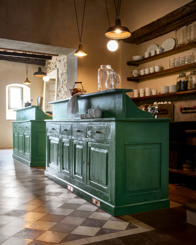Green wooden bar with cabinets, surrounded by shelves holding glassware, in a rustic kitchen setting.